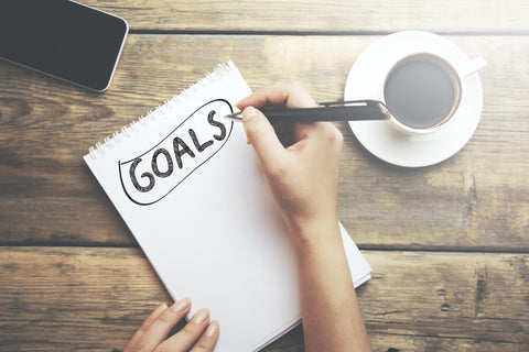 A wooden table with a white notepad, with a person's hand writing "GOALS" on it. To their right is a cup of coffee. 